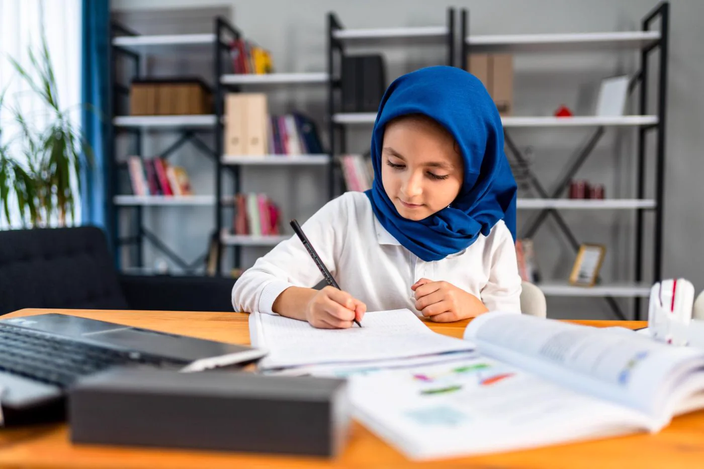 Girl in a blue hijab writing in a notebook at her desk